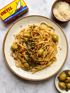 Plate of spaghetti with green olives and herbs beside a tin of Ortiz anchovies and grated cheese