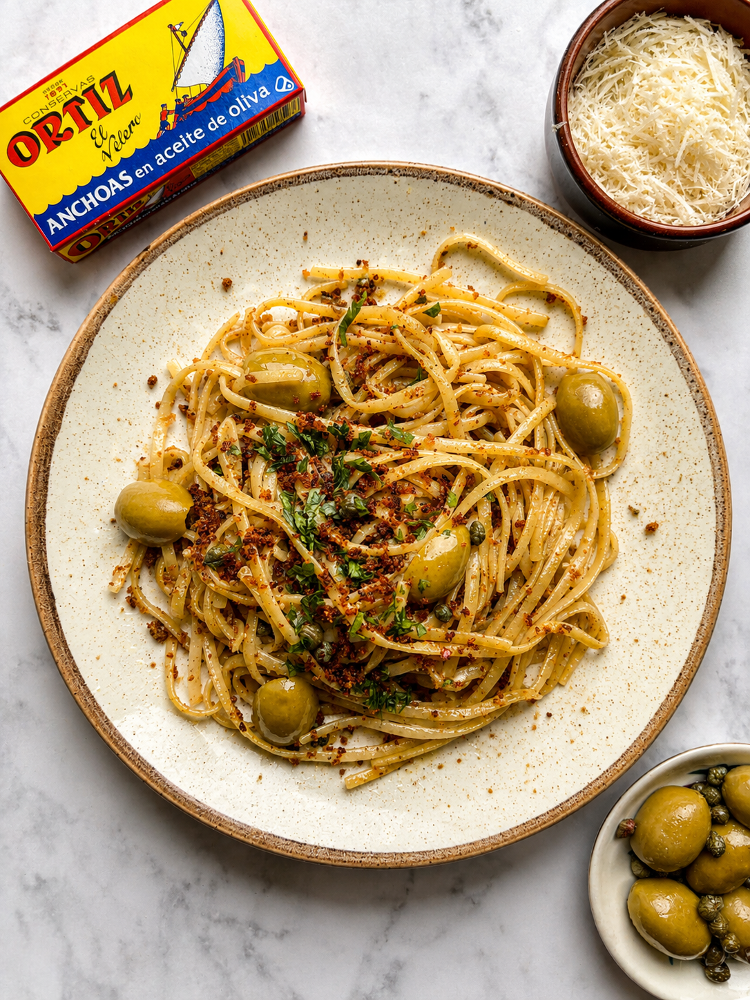 Plate of spaghetti with green olives and herbs beside a tin of Ortiz anchovies and grated cheese