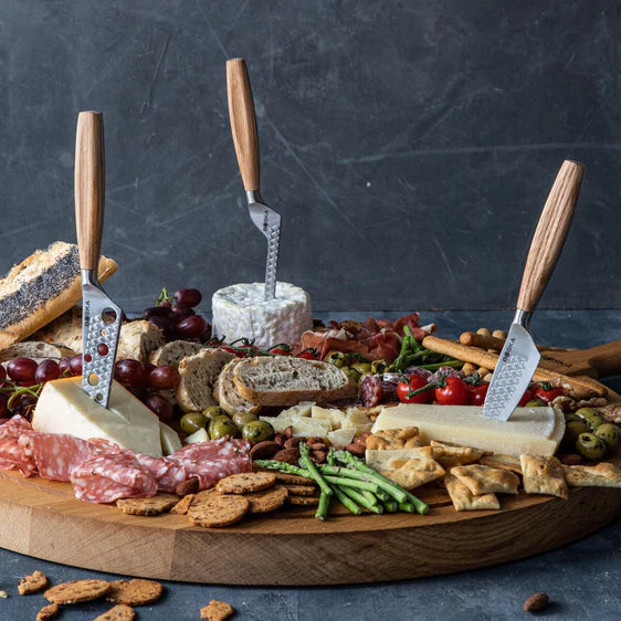 Assorted meats, cheeses, and crackers on a wooden board with cheese knives against a dark background.