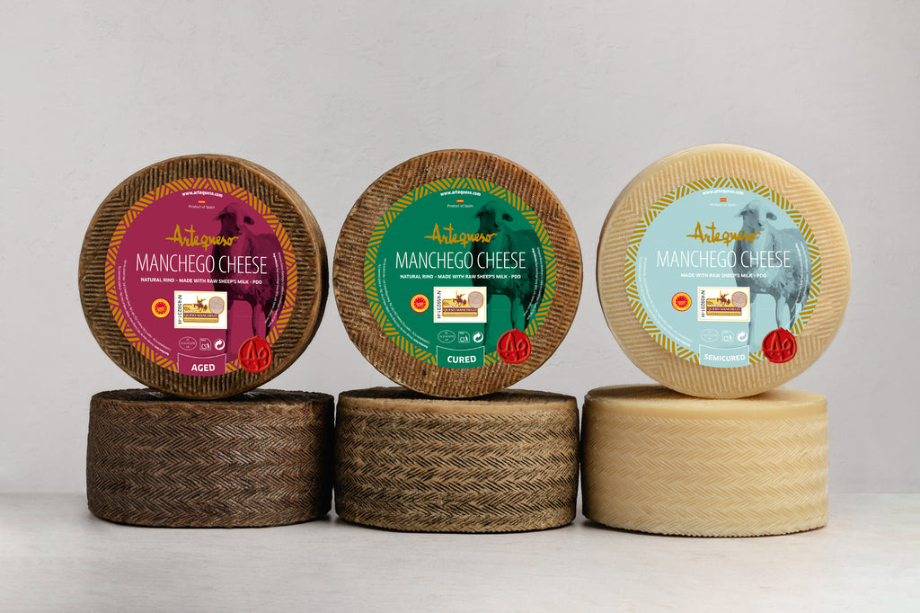 Three wheels of Manchego cheese with decorative labels on a light gray background