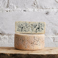 Two wheels of blue cheese stacked on a wooden surface with a white stone background