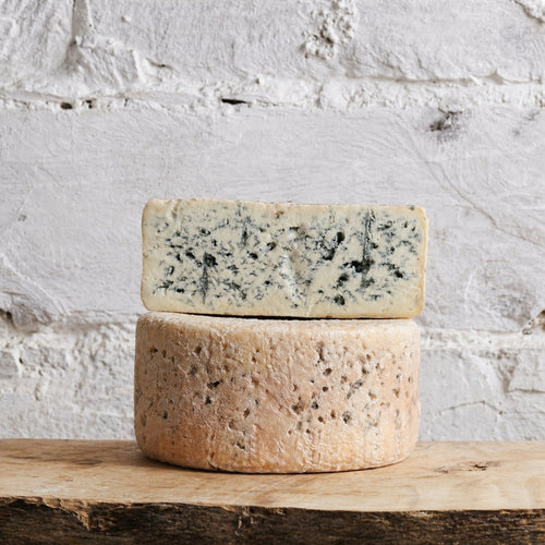 Two wheels of blue cheese stacked on a wooden surface with a white stone background