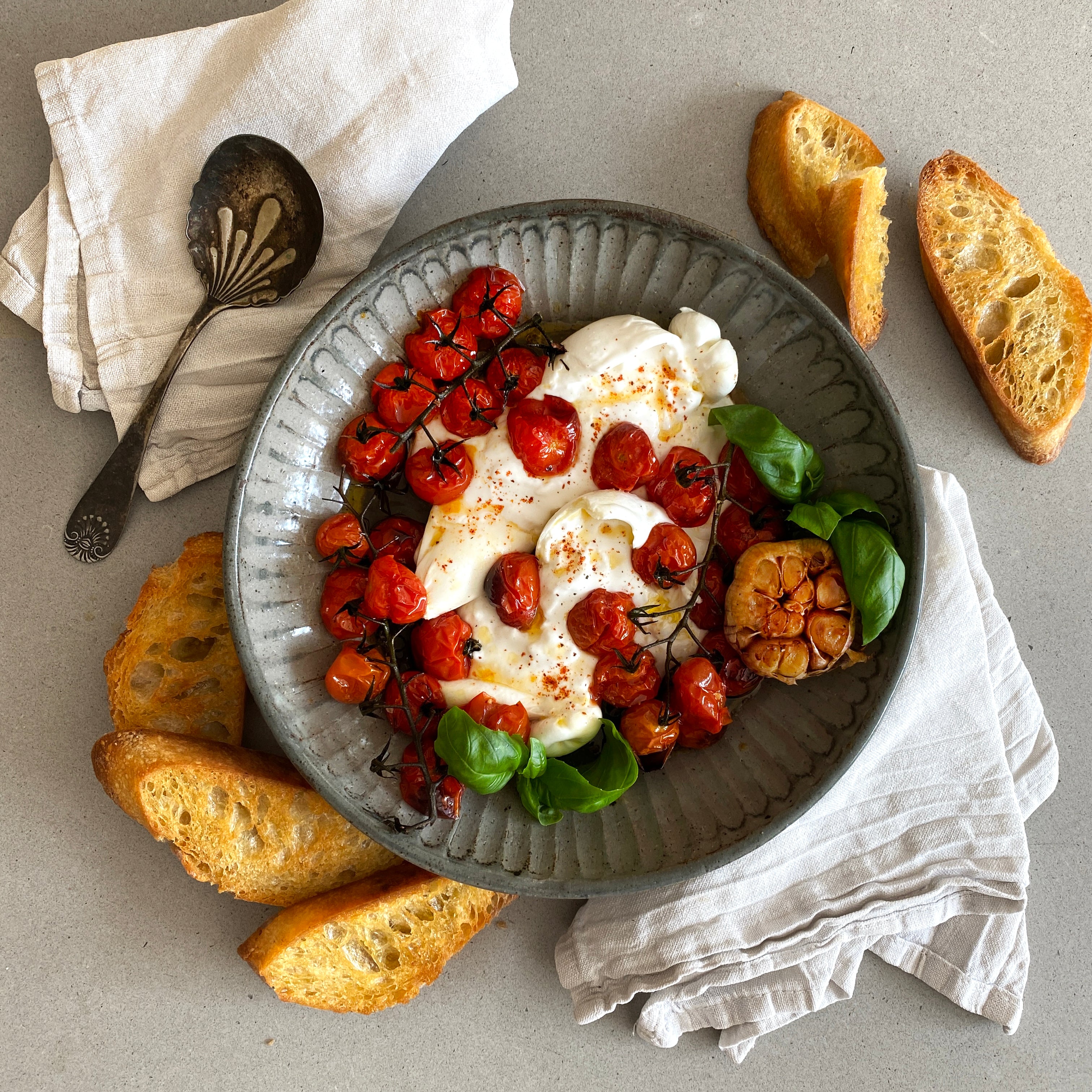 Gray bowl with roasted tomatoes and cheese, surrounded by bread slices on a light gray surface.