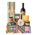 A curated gourmet gift hamper arranged in a colorful patterned box, featuring a bottle of red wine, a bottle of cashel blue, jars of spreads and preserves, crackers, and a boxed item, with a tall striped gift box behind it on a white background.