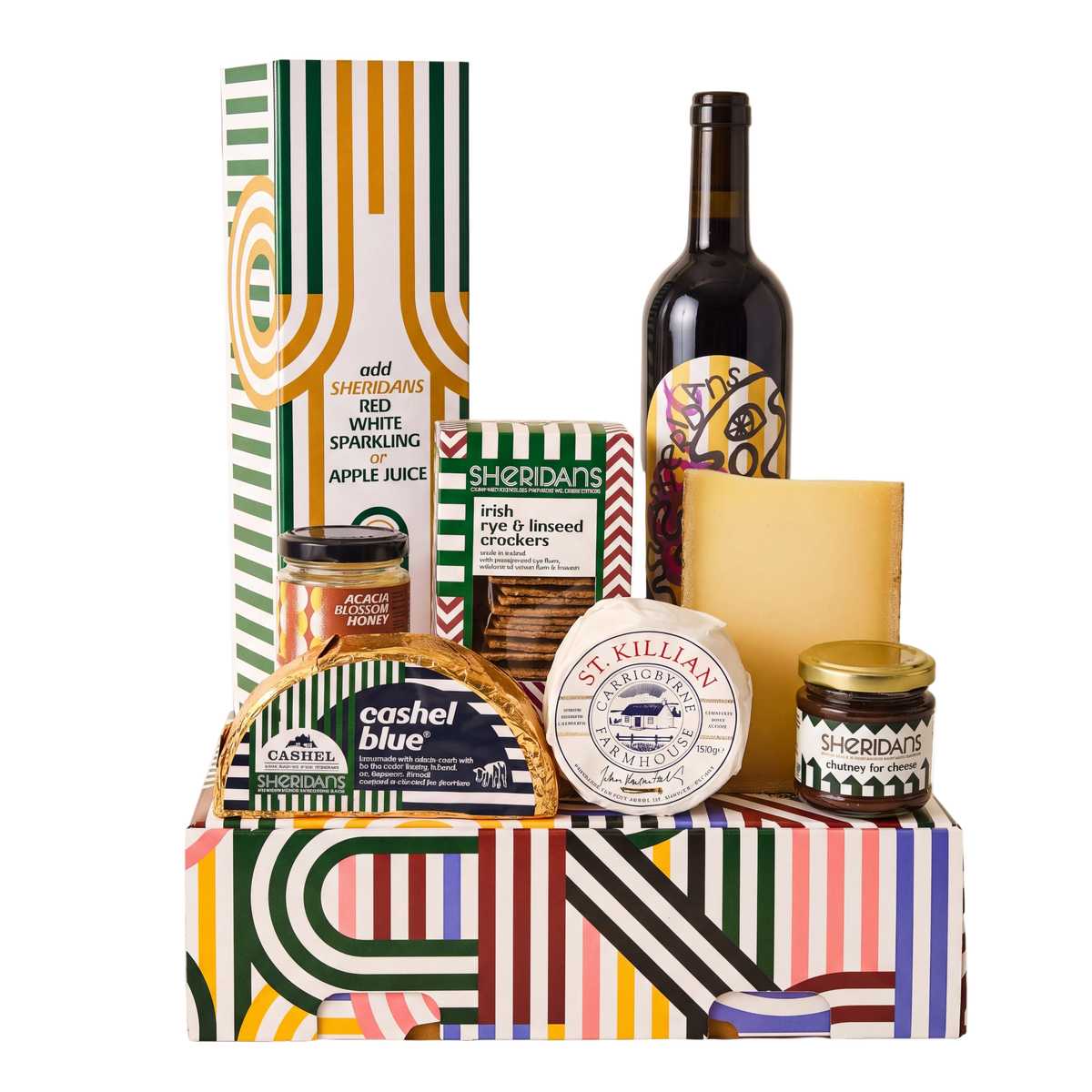 A curated gourmet gift hamper arranged in a colorful patterned box, featuring a bottle of red wine, a bottle of cashel blue, jars of spreads and preserves, crackers, and a boxed item, with a tall striped gift box behind it on a white background.