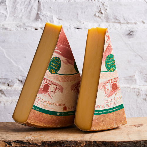 Two wedges of Comte Fort Saint Antoine cheese on a wooden surface with a white stone background.