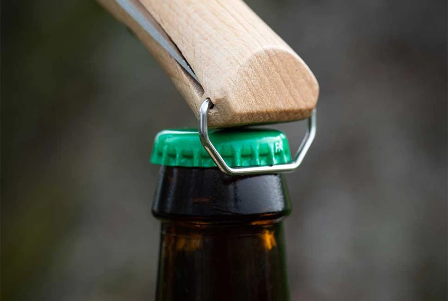 Close-up of a wooden bottle opener with a metal loop resting on a green bottle cap, with part of a dark brown glass bottle visible against a blurred gray background.