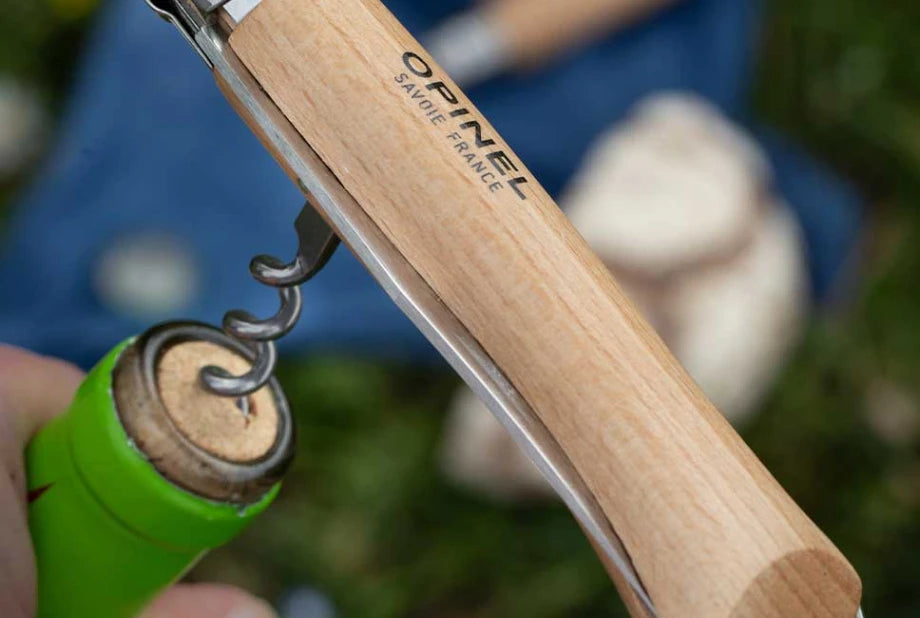 A close-up of a wooden corkscrew opener with the OFINEL brand name printed on it, being used to remove a cork from a green bottle. The background is a soft-focus outdoor scene with blue fabric and green grass.