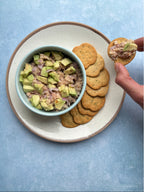 Bowl of tuna salad with avocado on a plate with crackers, hand dipping a cracker into the salad.