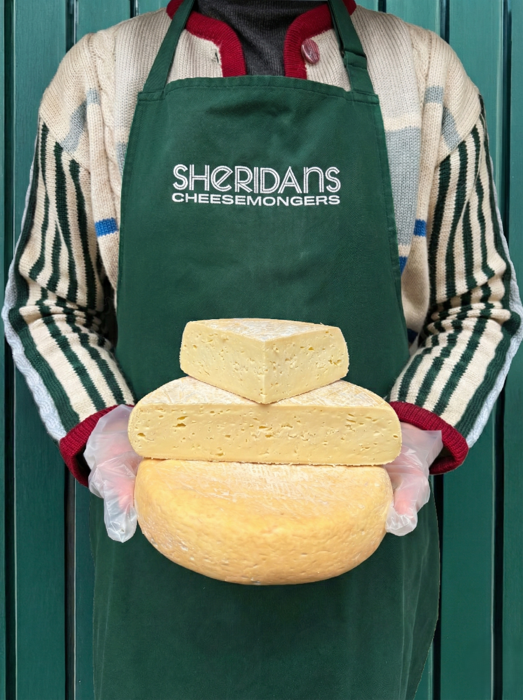 Person wearing a green 'Sheridan's Cheesemongers' apron holding a stack of cheeses.