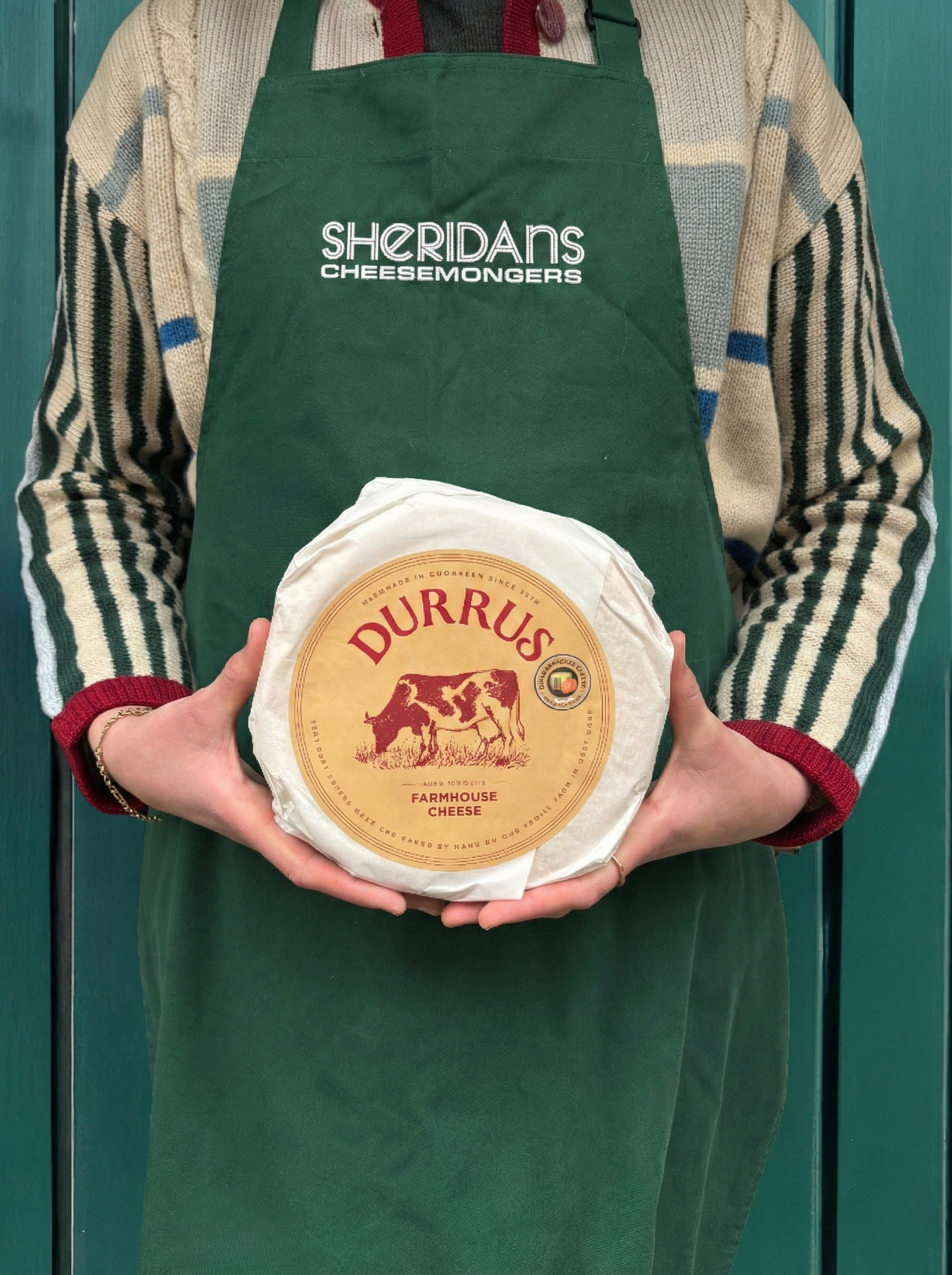 Person wearing a green apron with 'Sheridan's Cheesemongers' holding a Durrus Farmhouse Cheese.