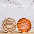 Two round cheese wheels with wooden rinds on a light wooden surface against a white marble background.