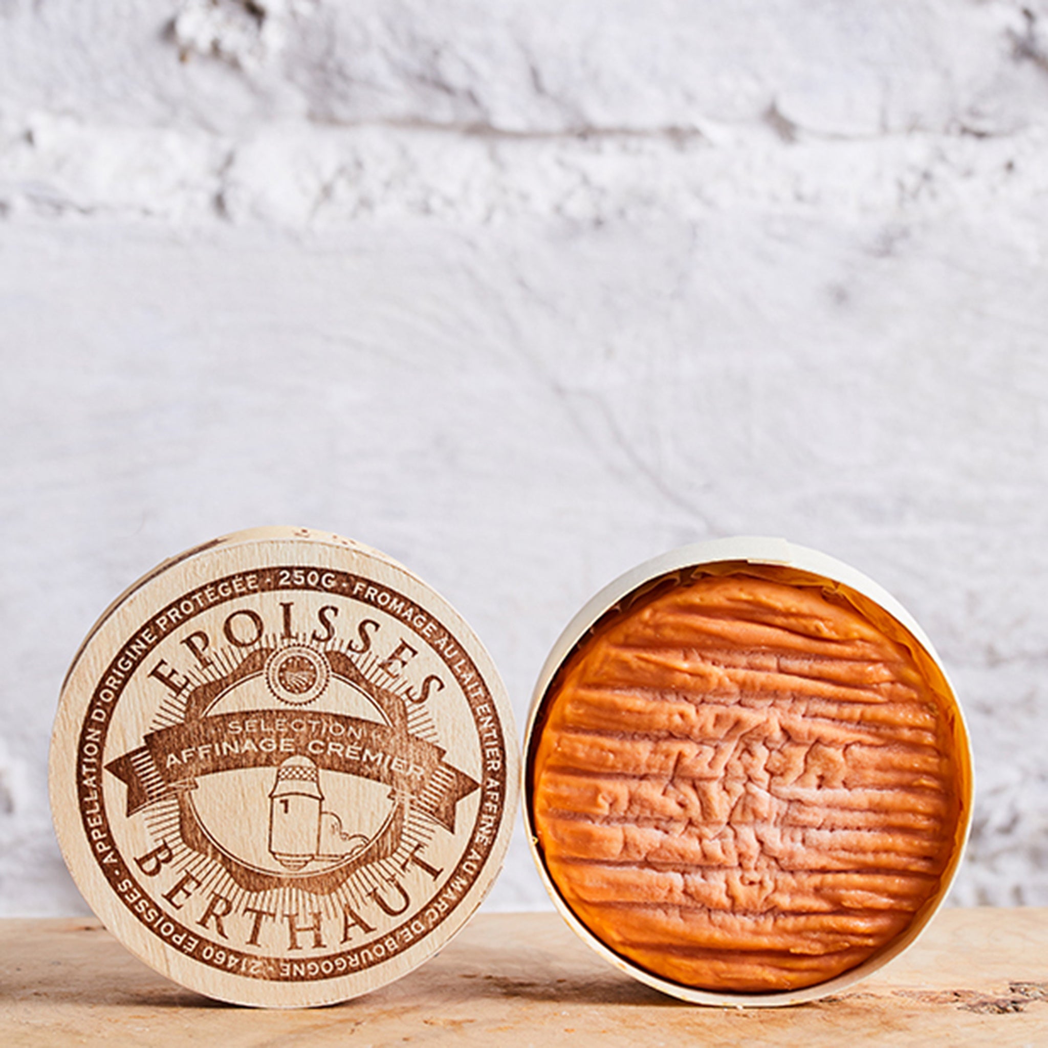 Two round cheese wheels with wooden rinds on a light wooden surface against a white marble background.