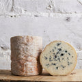 Two types of cheese, a whole and a sliced one, on a wooden board with a light gray textured background.