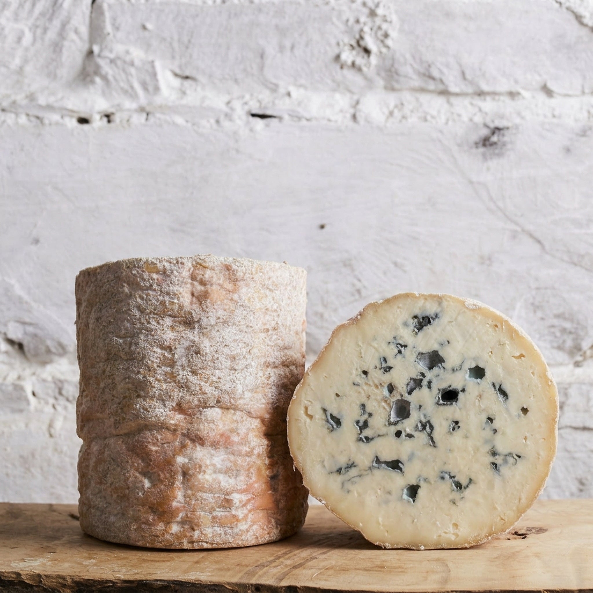 Two types of cheese, a whole and a sliced one, on a wooden board with a light gray textured background.