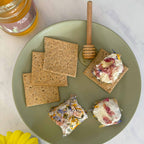 Plate with crackers topped with a spread, a honey dipper, and a jar of honey on a marble surface with yellow flowers.