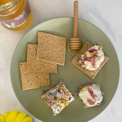 Plate with crackers topped with a spread, a honey dipper, and a jar of honey on a marble surface with yellow flowers.
