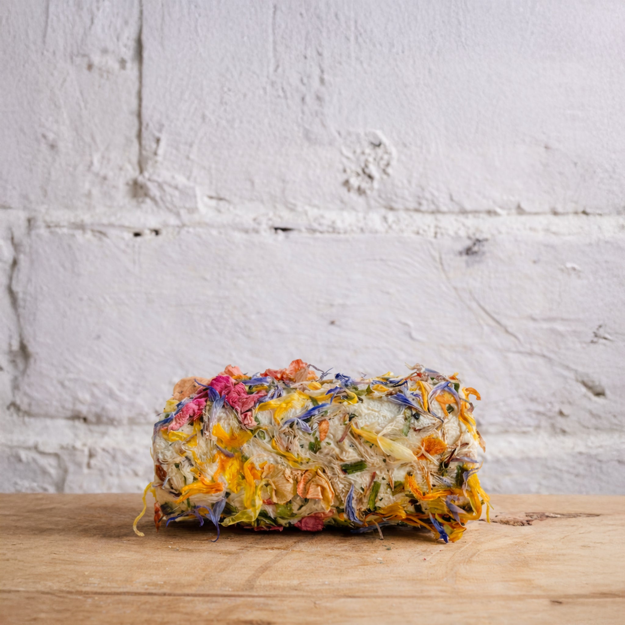 Loaf of bread with colorful toppings on a wooden surface against a white brick wall.