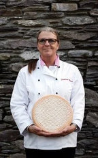 Person wearing a chef's coat holding a round object against a stone wall.