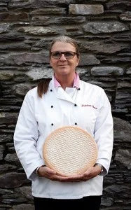 Person wearing a chef's coat holding a round object against a stone wall.