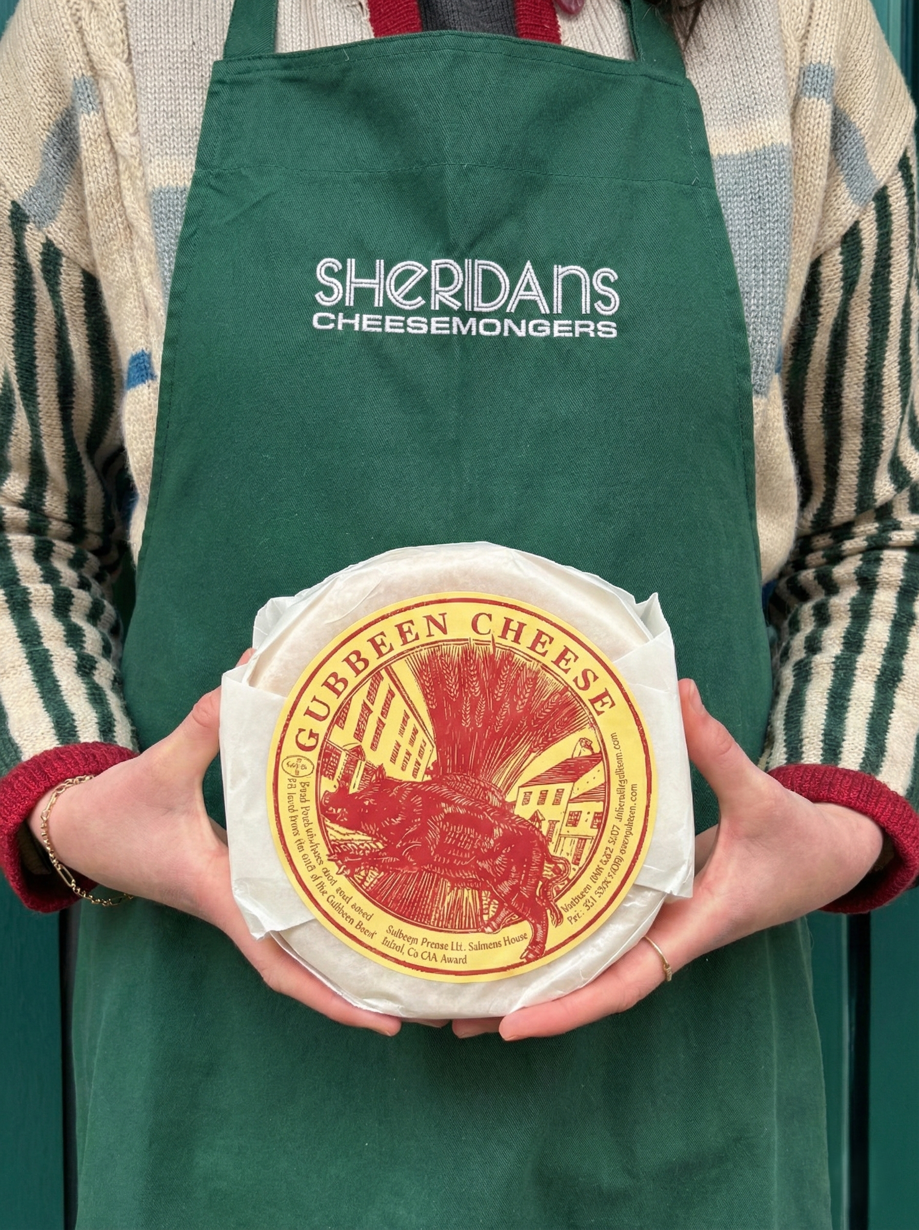 Person wearing a green apron with 'Sheridan's Cheesemongers' holding a Gubbeen Cheese.