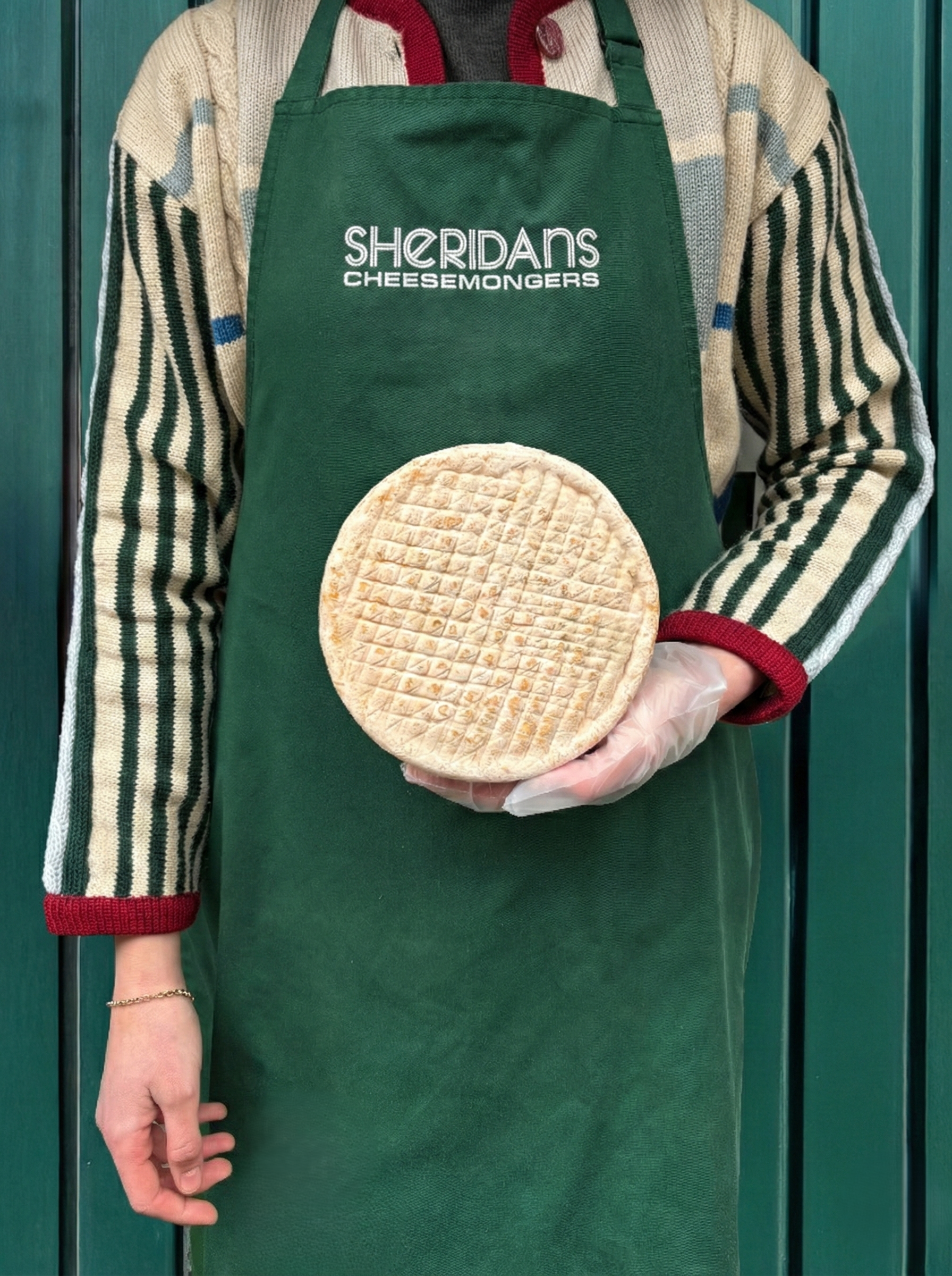 Person wearing a green apron with 'Sheridan's Cheesemongers' holding a wheel of cheese against a green background.
