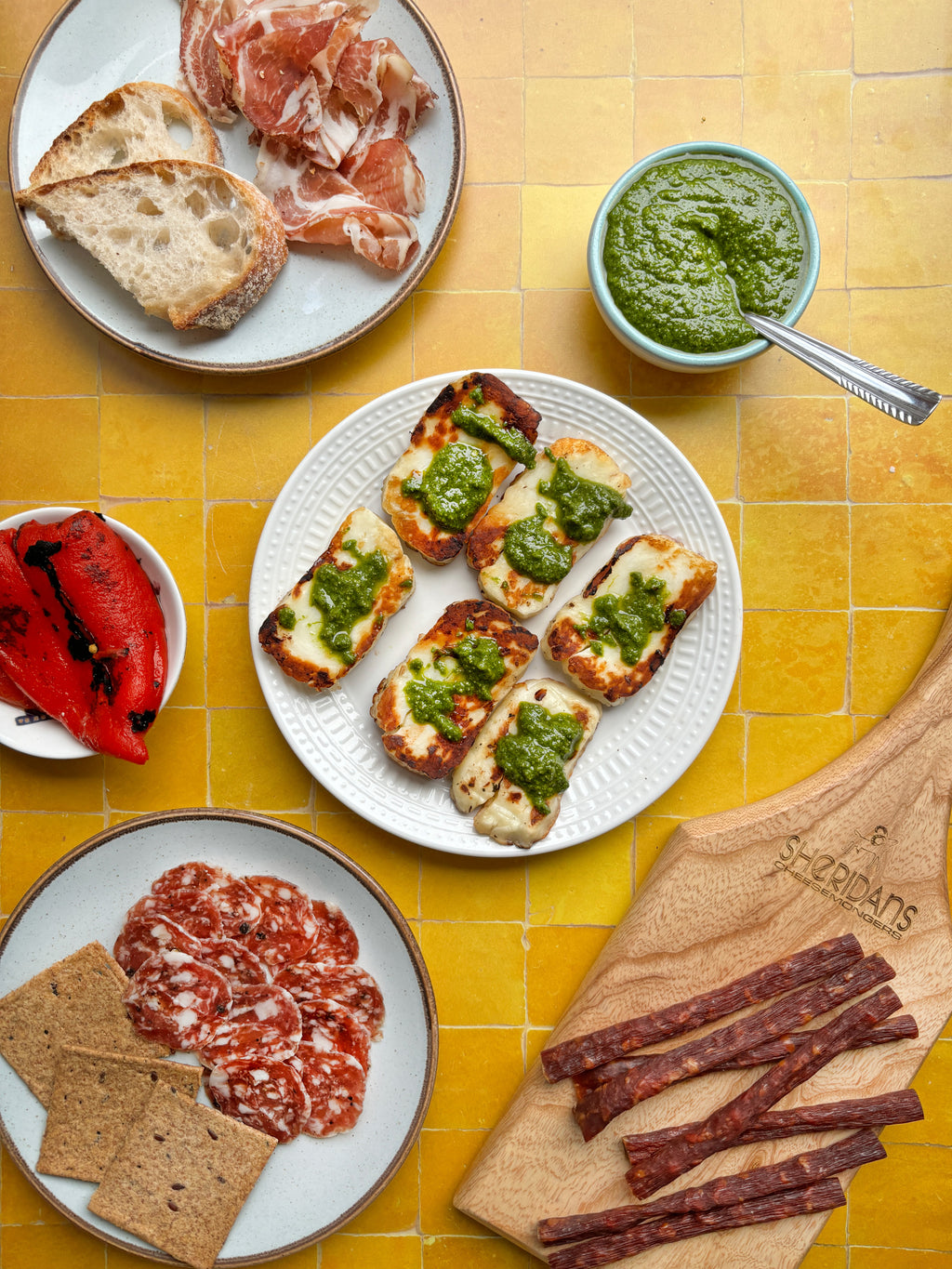 Plates of food including bread, prosciutto, pesto, and salami on a yellow tiled table.
