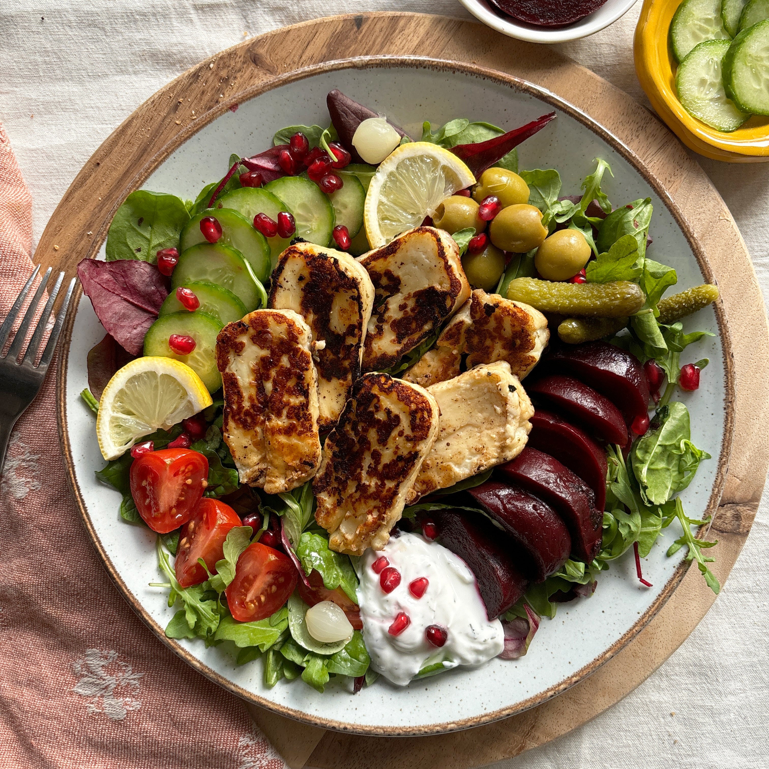Plated salad with grilled chicken, vegetables, and a side of pomegranate seeds on a wooden board.