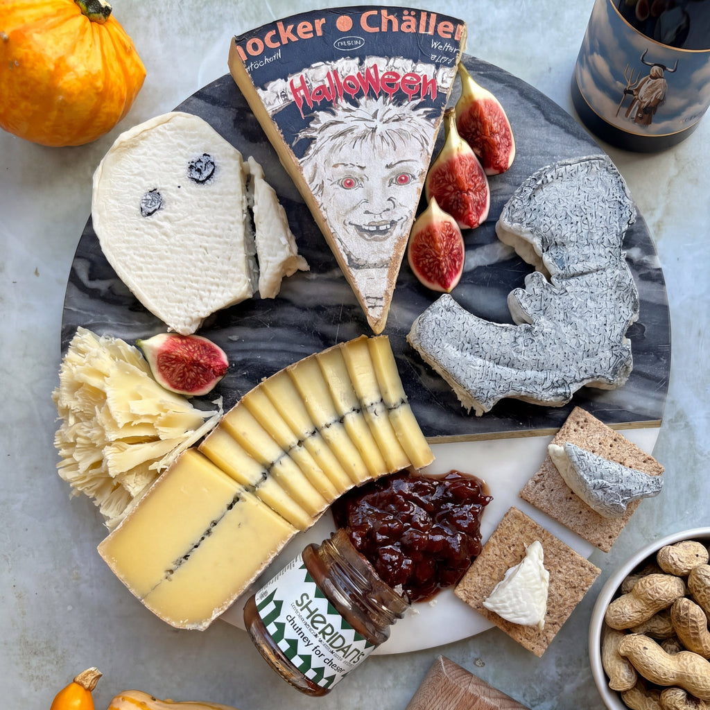 A styled Halloween-themed cheese board arrangement on a marble platter, featuring assorted cheeses, figs, crackers, peanut butter, and seasonal decor. Visible cheese labels include 'Hucker Chäller' and 'Schön's'.