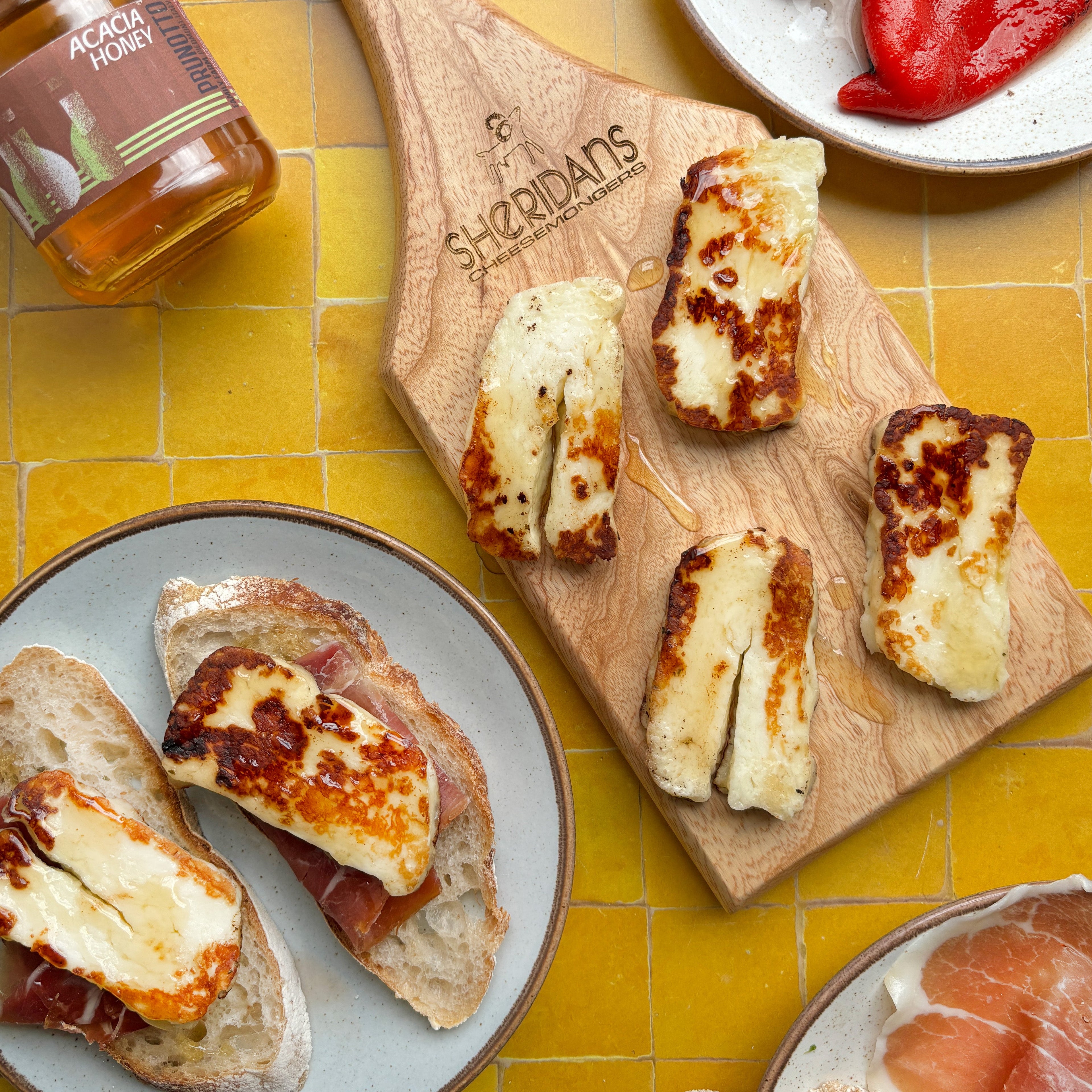 Wooden cutting board with grilled cheese and prosciutto, jar of honey, and red peppers on a yellow tiled surface.