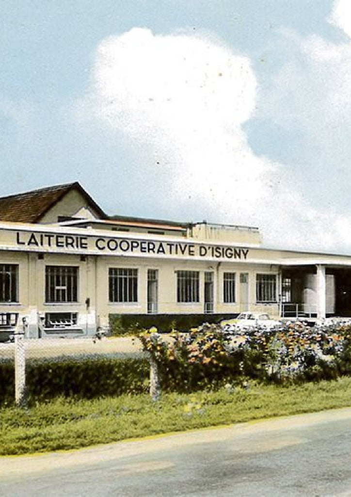 A vintage postcard or scanned photograph showing the exterior of a building labeled 'LAITERIE COOPERATIVE D'ISIGNY' under a blue sky with a large white cloud. The building is cream-colored with dark windows, and there are green hedges and flowers in the foreground.