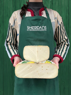 Person wearing a green apron with 'Sheridan's Cheesemongers' holding two blocks of cheese against a green background