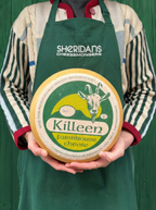 Person wearing a green apron with 'Sheridan's Cheesemongers' holding a Killeen Farmhouse Cheese wheel.