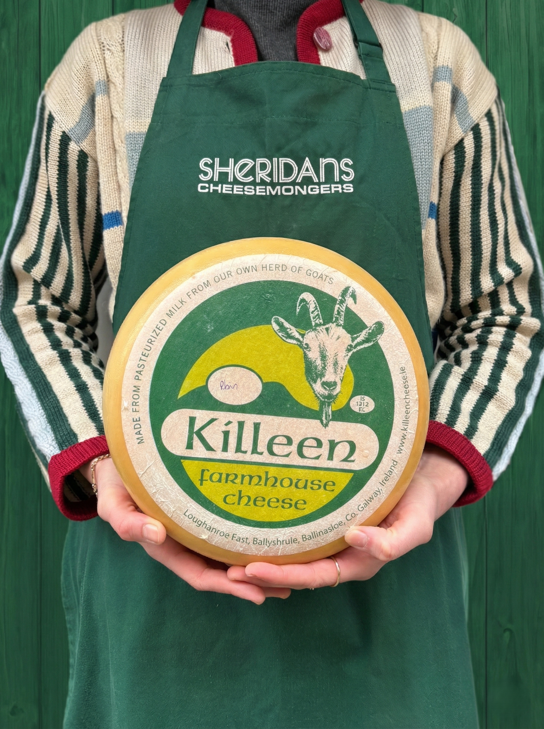 Person wearing a green apron with 'Sheridan's Cheesemongers' holding a Killeen Farmhouse Cheese wheel.