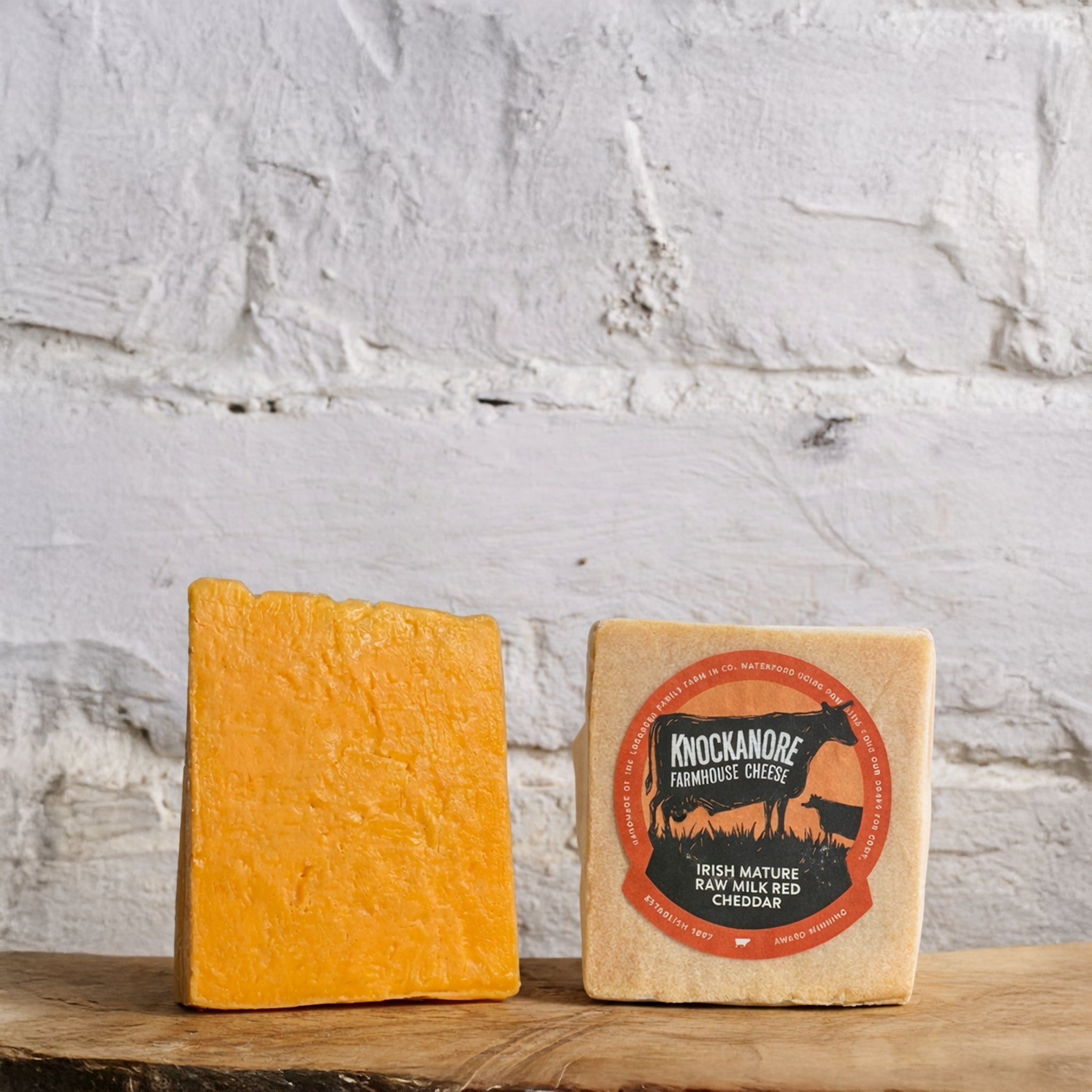 Two blocks of cheese with a label on a wooden surface against a white brick wall.
