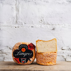 Langres cheese with packaging on a wooden surface against a white brick wall.