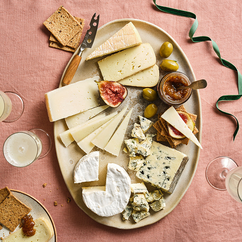 Assorted cheeses on a platter with figs, olives, and bread on a pink tablecloth.