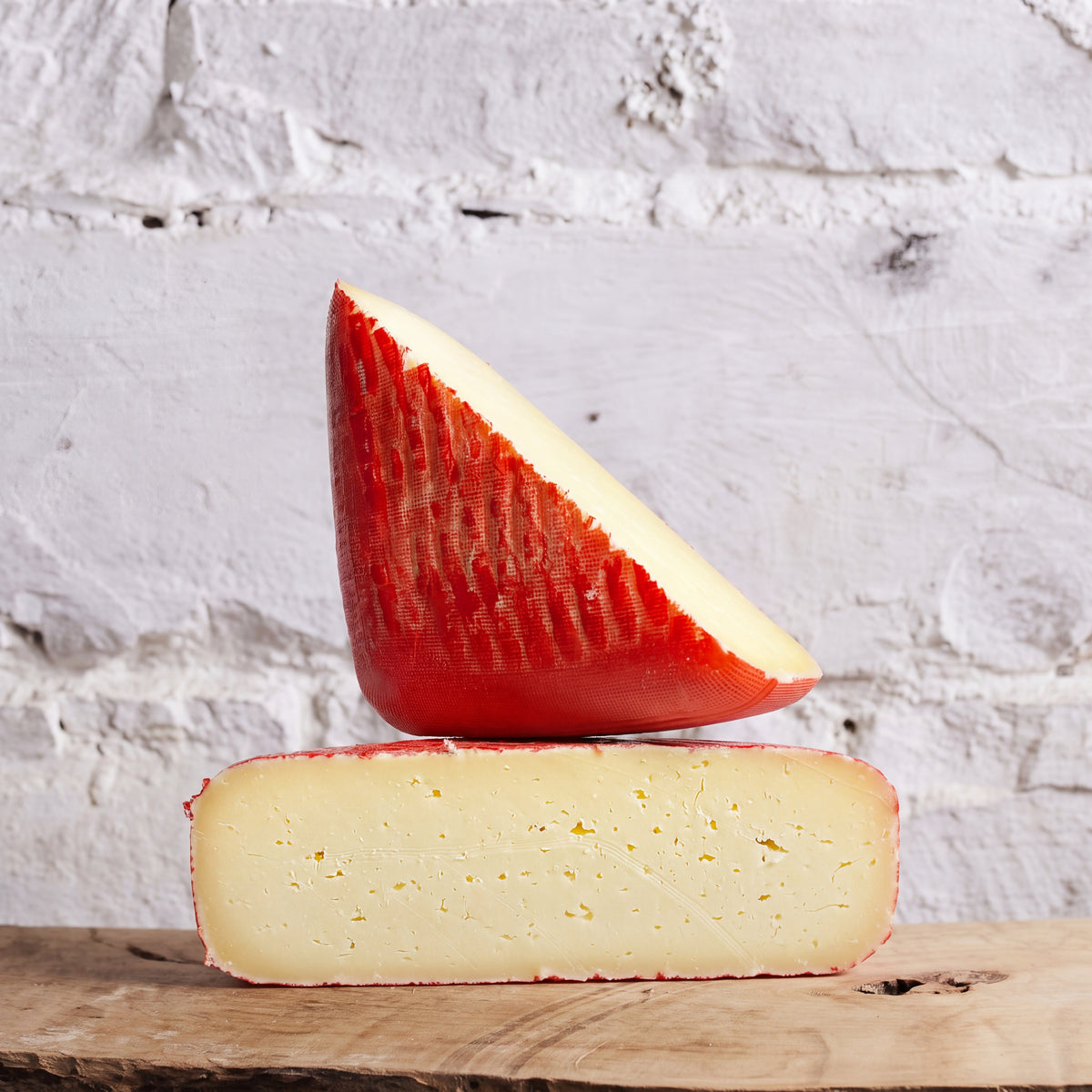 A wedge of red-skinned cheese resting on a rectangular block of the same cheese, displayed on a wooden surface against a white painted brick wall.