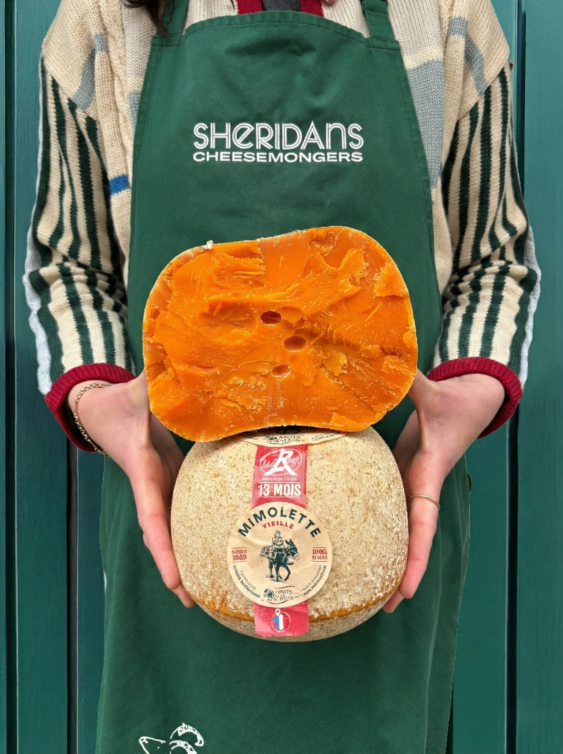Person wearing a Sheridan's Cheesemongers apron holding two types of cheese.
