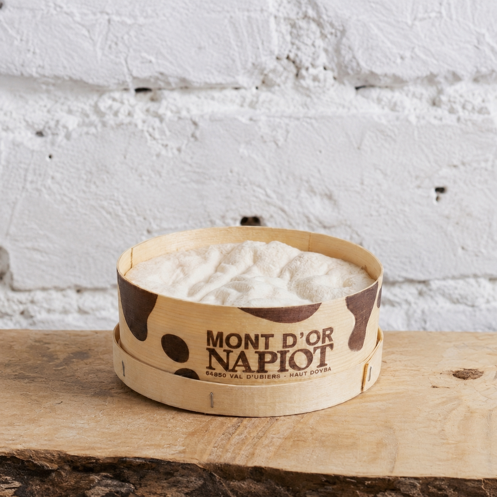 Wooden bowl with white product labeled 'Mont d'Or Napiot' on a rustic wooden surface with a white textured wall background.