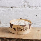 Wooden bowl with white product labeled 'Mont d'Or Napiot' on a rustic wooden surface with a white textured wall background.