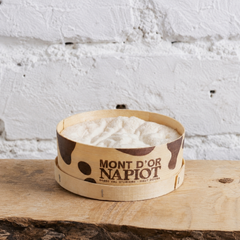 Wooden bowl with white product labeled 'Mont d'Or Napiot' on a rustic wooden surface with a white textured wall background.