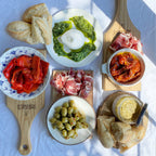 Assorted Italian antipasto including bread, olives, prosciutto, and pesto on a white surface.