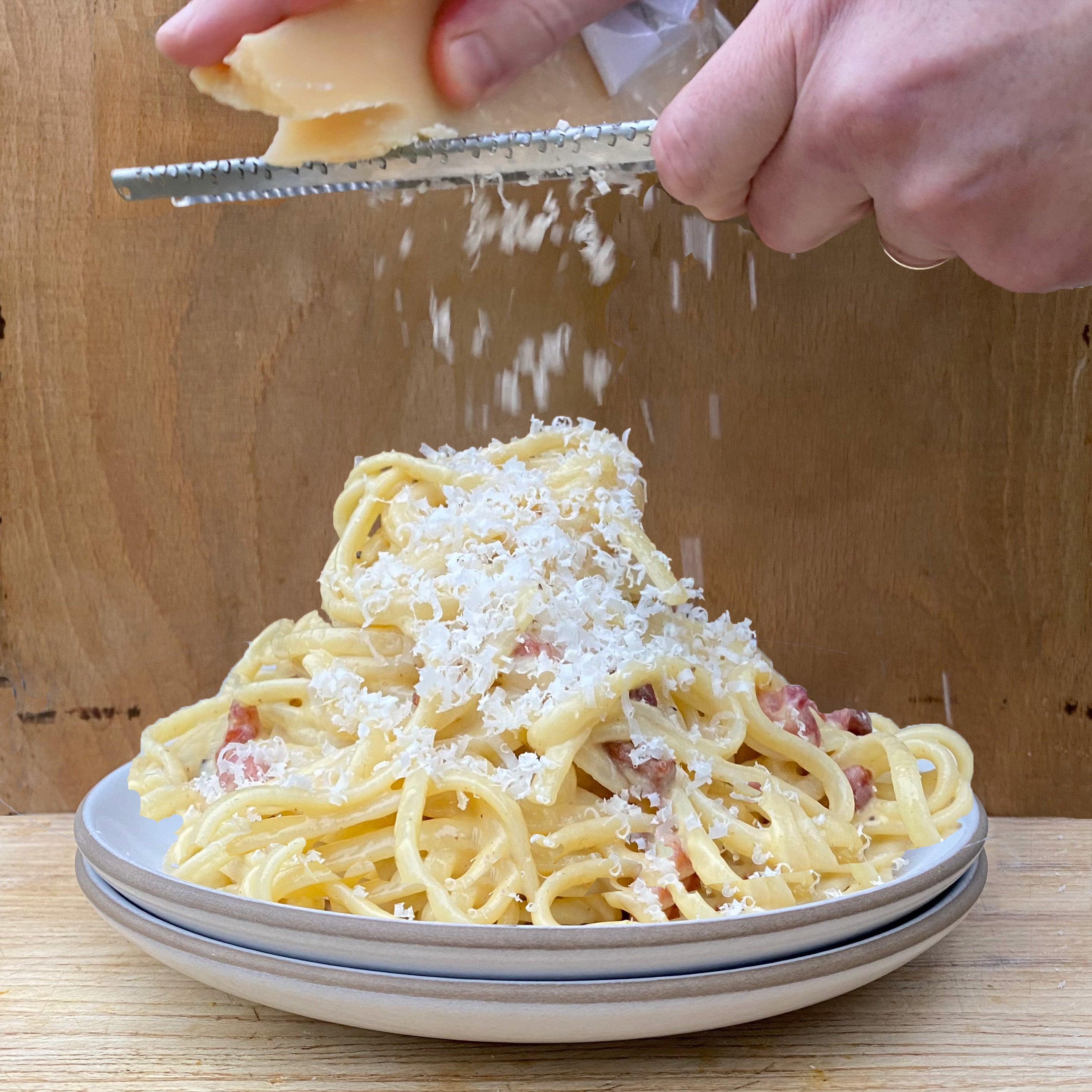 Person grating parmesan cheese onto a plate of spaghetti carbonara.