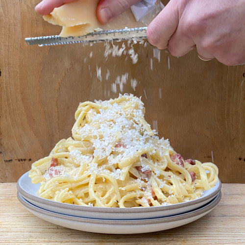 Person grating parmesan cheese onto a plate of spaghetti carbonara.