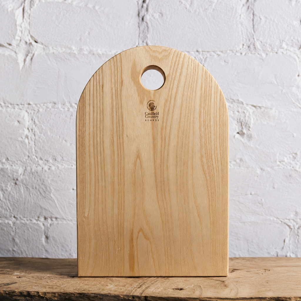 A light natural wood cutting board with a rounded top and a circular handle cutout, standing upright on a rustic wooden surface against a white painted brick wall. The brand name 'Cavelli' is printed near the top center.