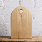 A light natural wood cutting board with a rounded top and a circular handle cutout, standing upright on a rustic wooden surface against a white painted brick wall. The brand name 'Cavelli' is printed near the top center.