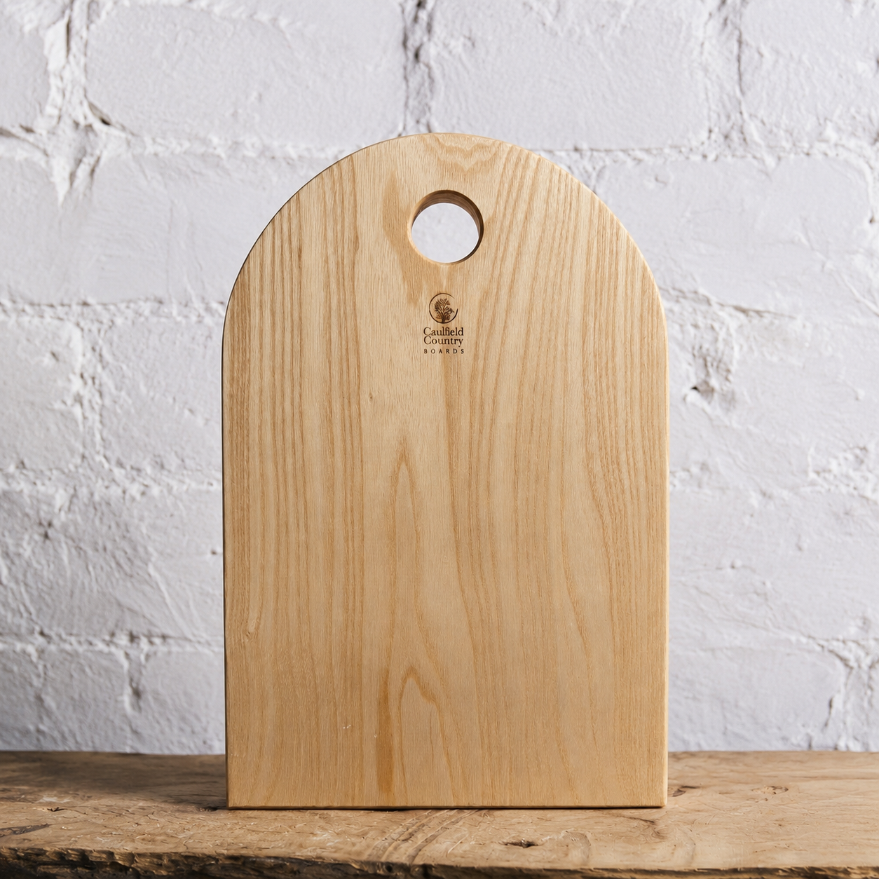 A light natural wood cutting board with a rounded top and a circular handle cutout, standing upright on a rustic wooden surface against a white painted brick wall. The brand name 'Cavelli' is printed near the top center.