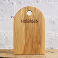 A natural wood cheese board with a rounded top and a circular handle cutout, featuring the engraved brand name 'SHERIDANS CHEESEMONGERS'. The board is standing upright on a wooden surface against a white painted brick wall.