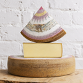 A wedge of artisanal cheese with a decorative printed paper cap and a thick rind, displayed on top of a cut wedge of the same cheese resting on a round wooden wheel, against a white brick wall background.